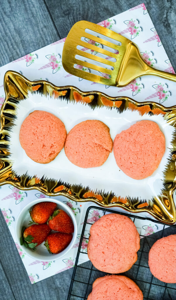 overhead view of 3 strawberry cake mix cookies on a platter next to a gold spatula and a bowl of strawberries