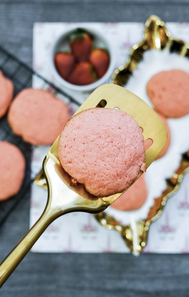 closeup of a strawberry cake mix cookie on a gold spatula with more cookies and strawberries in the background