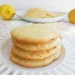 a stack of lemon cake cookies on a white plate with more cookies and lemons in the background