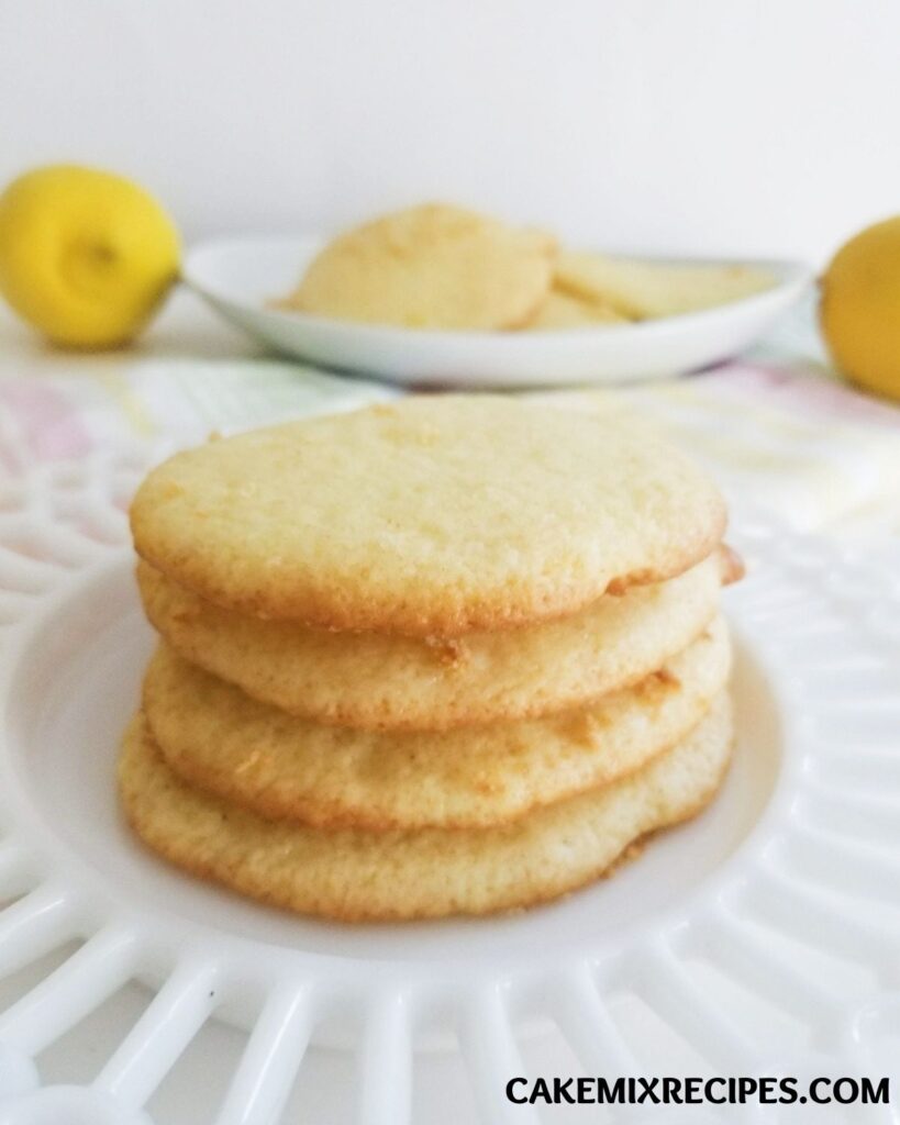 a stack of lemon cake cookies on a white plate with more cookies and lemons blurred in the background