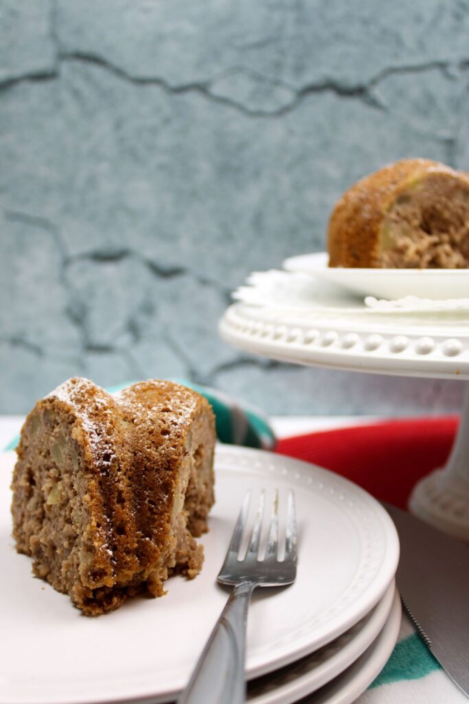 a slice of applesauce bundt cake next to a fork on a white plate with more cake on a white cake platter in the background