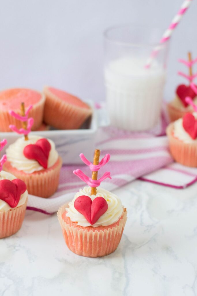 a Cupid's bow valentine cupcake in the center of the image with more decorated cupcakes around it, and more unfrosted cupcakes and a glass of milk in the background