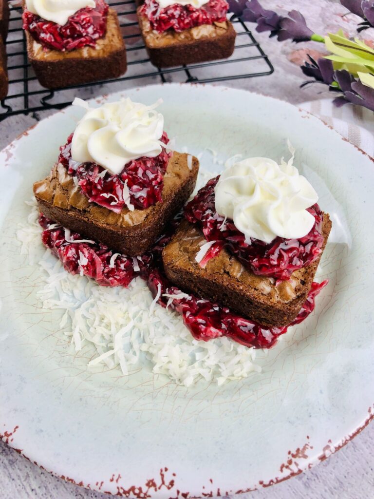 two brownies with raspberry lamington on a white plate topped with whipped cream and surrounded by shredded coconut