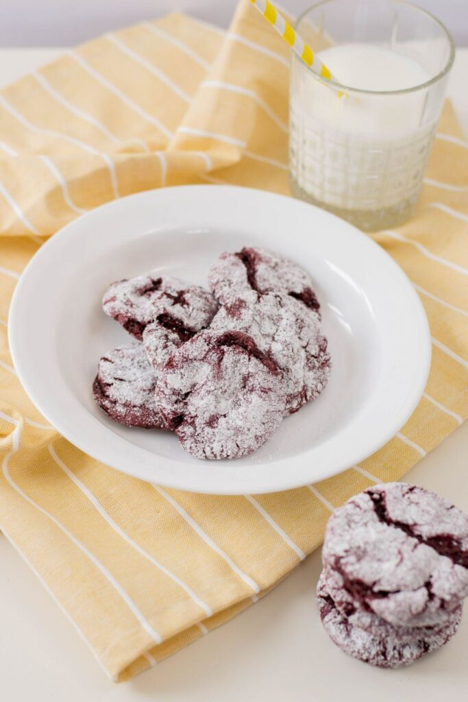 red velvet cake cookies on a white plate on a yellow and white striped cloth next to a glass of milk and more stacked cookies