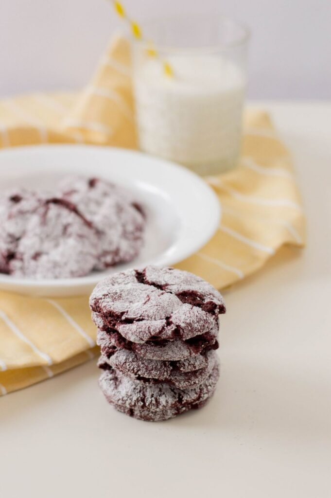 a stack of red velvet cake cookies with more cookies on a white plate on a yellow and white striped cloth next to a glass of milk in the background