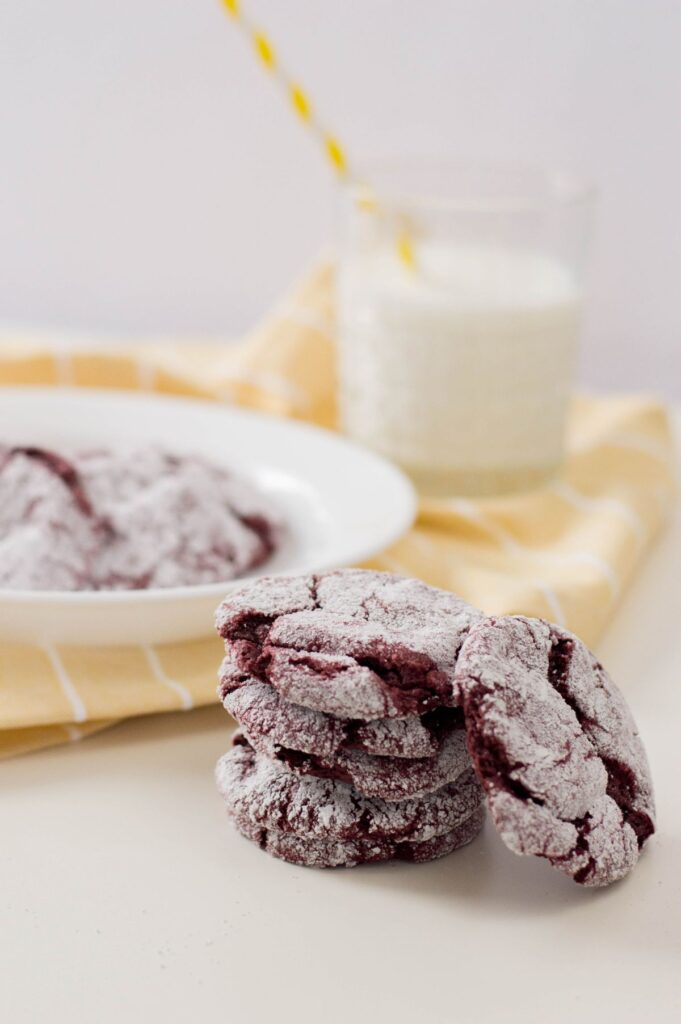 a stack of red velvet cake cookies in front of more cookies on a plate next to a glass of milk on a yellow and white striped cloth