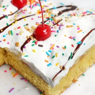 banana split poke cake topped with frosting, rainbow sprinkles, chocolate drizzle, and cherries