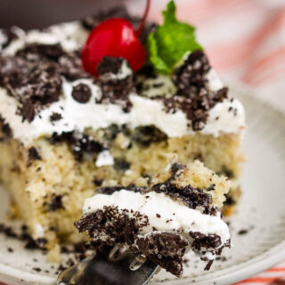 oreo poke cake topped with vanilla frosting, oreo pieces, and a cherry