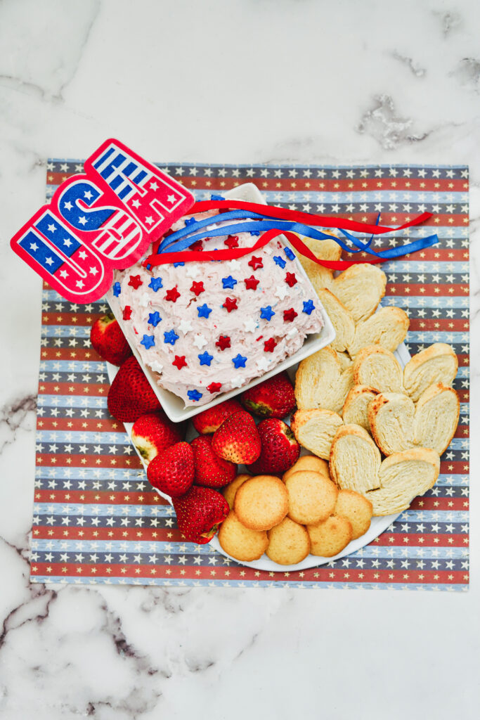 strawberries and cookies next to patriotic dunkaroo dip in a white dish next to a USA sign and on a red, white and blue striped cloth
