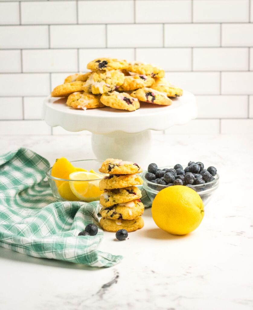 a stack of Lemon Blueberry Cookies on a white cake plate next to more cookies, lemons, blueberries and a green and white cloth