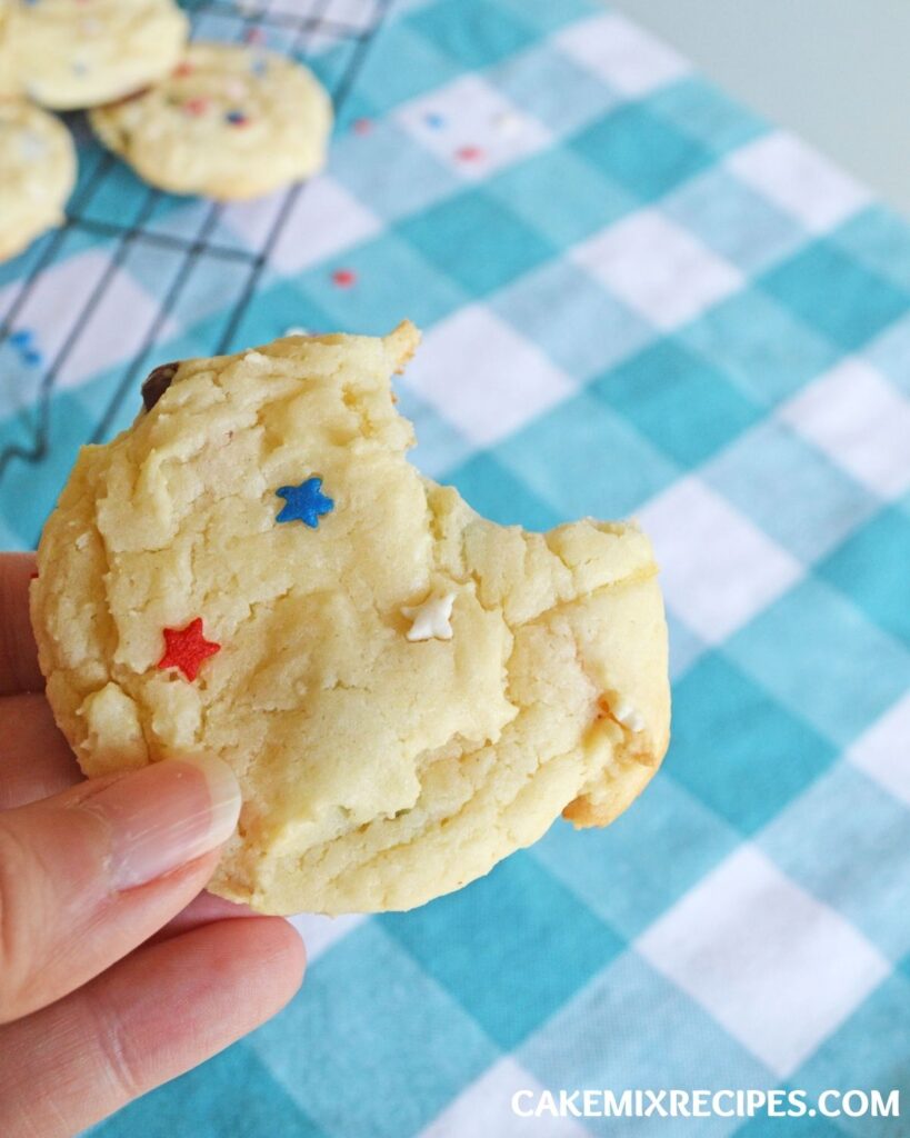 a hand holding a cookie with a bite taken out of it with the rest of the Chocolate Chip Cake Mix Cookies in the background on a wire rack on a blue and white checkered cloth