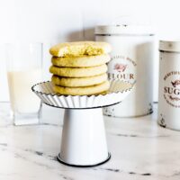 sugar cookies with cake mix stacked on a white cake stand next to a glass of milk, and flour and sugar canisters