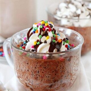 chocolate mug cake in a clear mug topped with whipped cream, chocolate syrup, and rainbow sprinkles