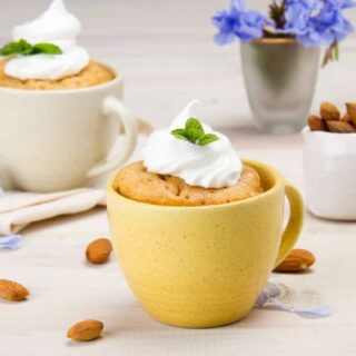two mugs of vanilla mug cake with cool whip on top and green leaf garnishes, surrounded by almonds