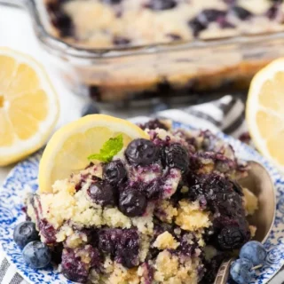 Small blue plate of lemon blueberry dump cake with spoon and lemon on side, with a larger casserole dish of dump cake in the background