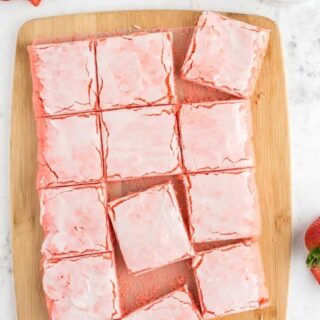 squares of pink strawberry brownies on a wooden cutting board with strawberries on the side