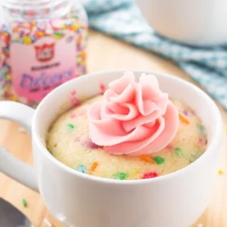 vanilla mug cake with rainbow sprinkles in it, in a white mug, with pink icing on top