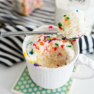 vanilla mug cake with rainbow sprinkles inside, in a white mug with a piece taken out on a spoon