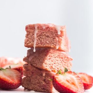 Strawberry brownie squares stacked on top of one another with cut strawberries around them and pink icing on top