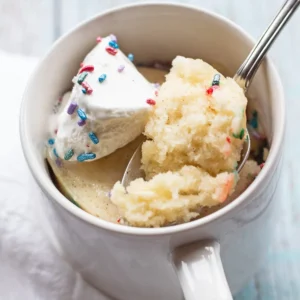 Vanilla mug cake with rainbow sprinkles and cool whip on top, in a white mug