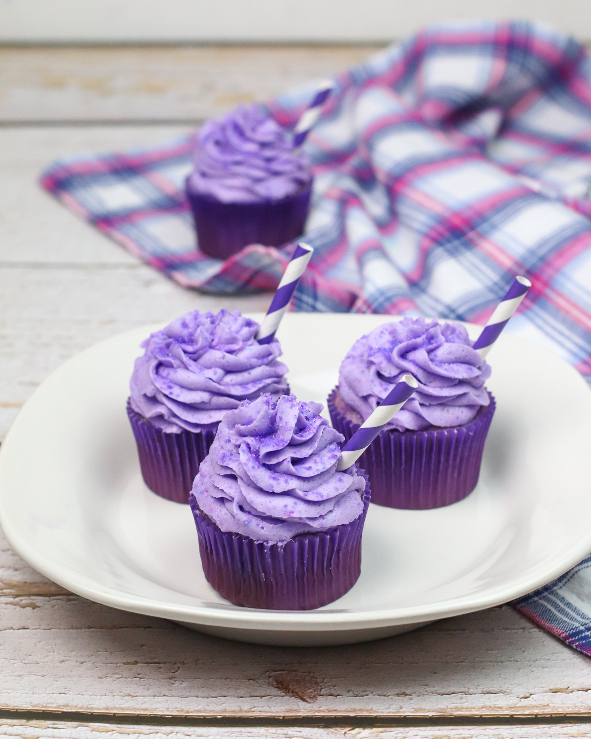 3 grape soda cupcakes on a white plate with another cupcake and a purple and white cloth in the background