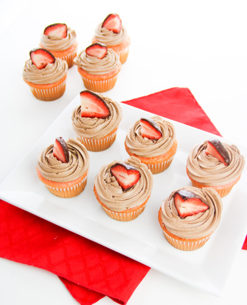 6 chocolate cupcakes with strawberry on top on a white platter on a red cloth with more cupcakes in the background
