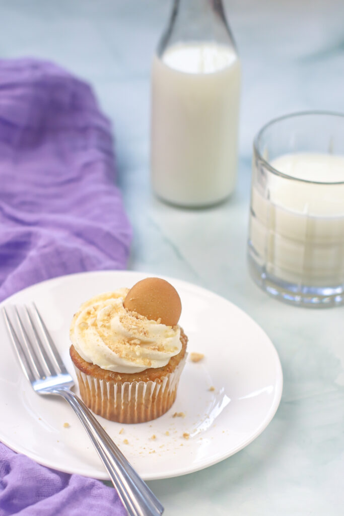 a banana pudding cupcake on a white plate next to a fork on a purple cloth next to a glass of milk and a glass container of milk