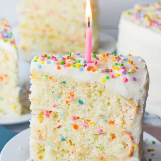 Big slice of vanilla funfetti cake with white icing, rainbow sprinkles and a pink candle on top