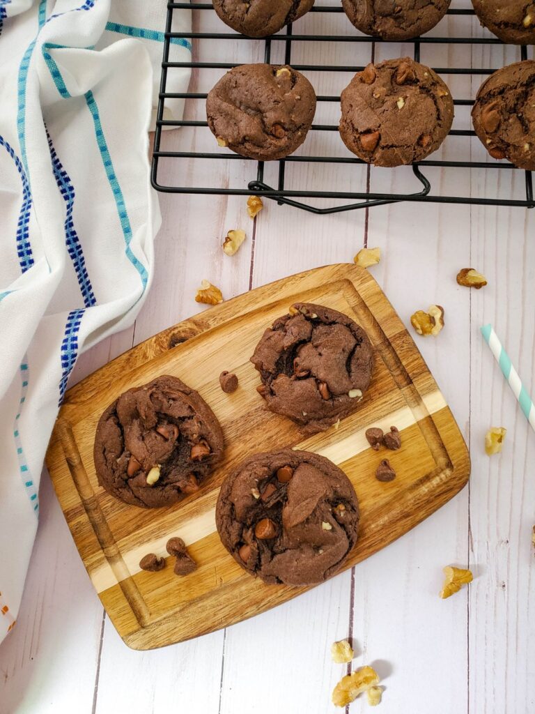 3 double chocolate chip walnut cookies on a wooden board next to more cookies on a wire rack