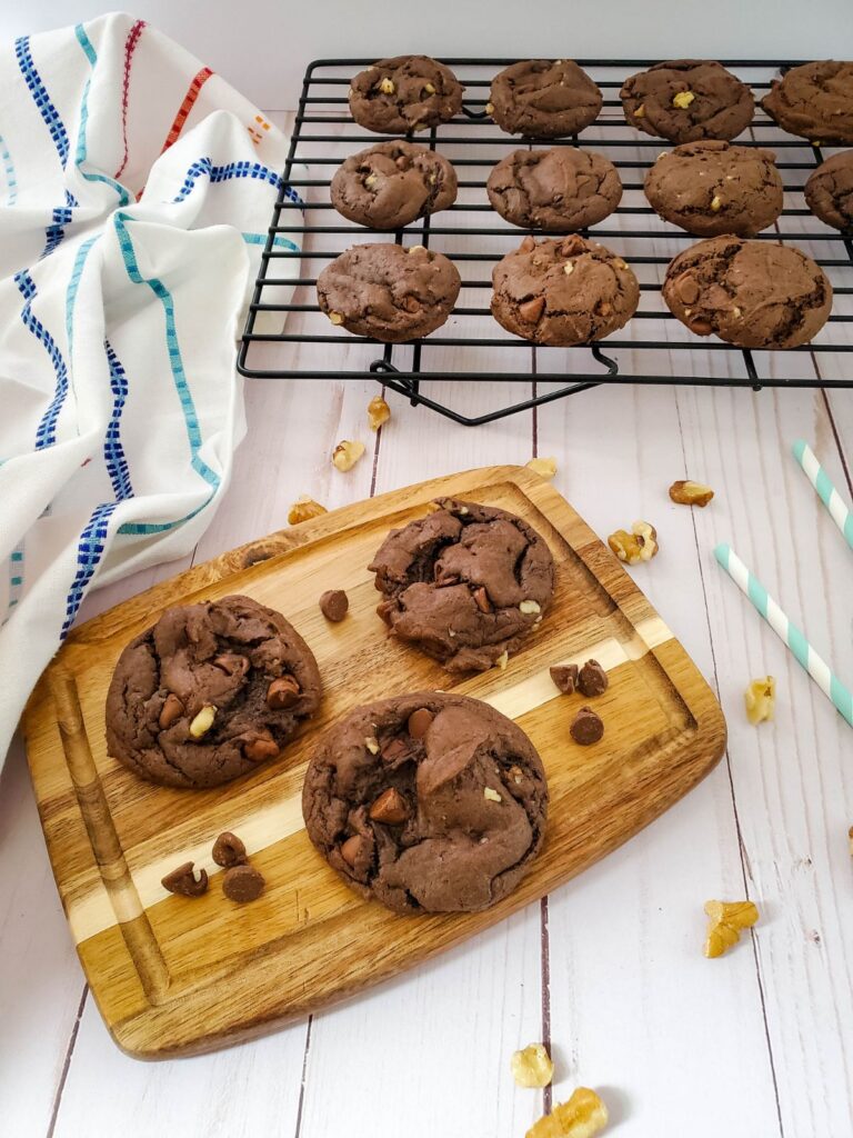 3 walnut and chocolate chip cookies on a wooden board next to more cookies on a wire rack