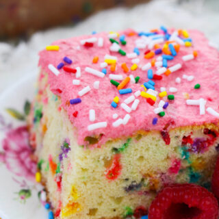 piece of vanilla funfetti cake with pink icing and rainbow sprinkles on top, with a raspberry on the side
