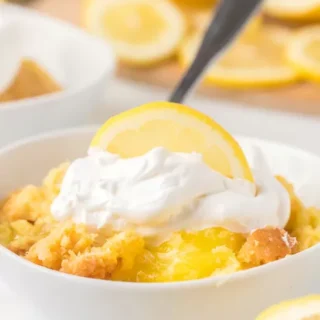 white bowl of lemon dump cake with fresh cut lemons and cool whip on top