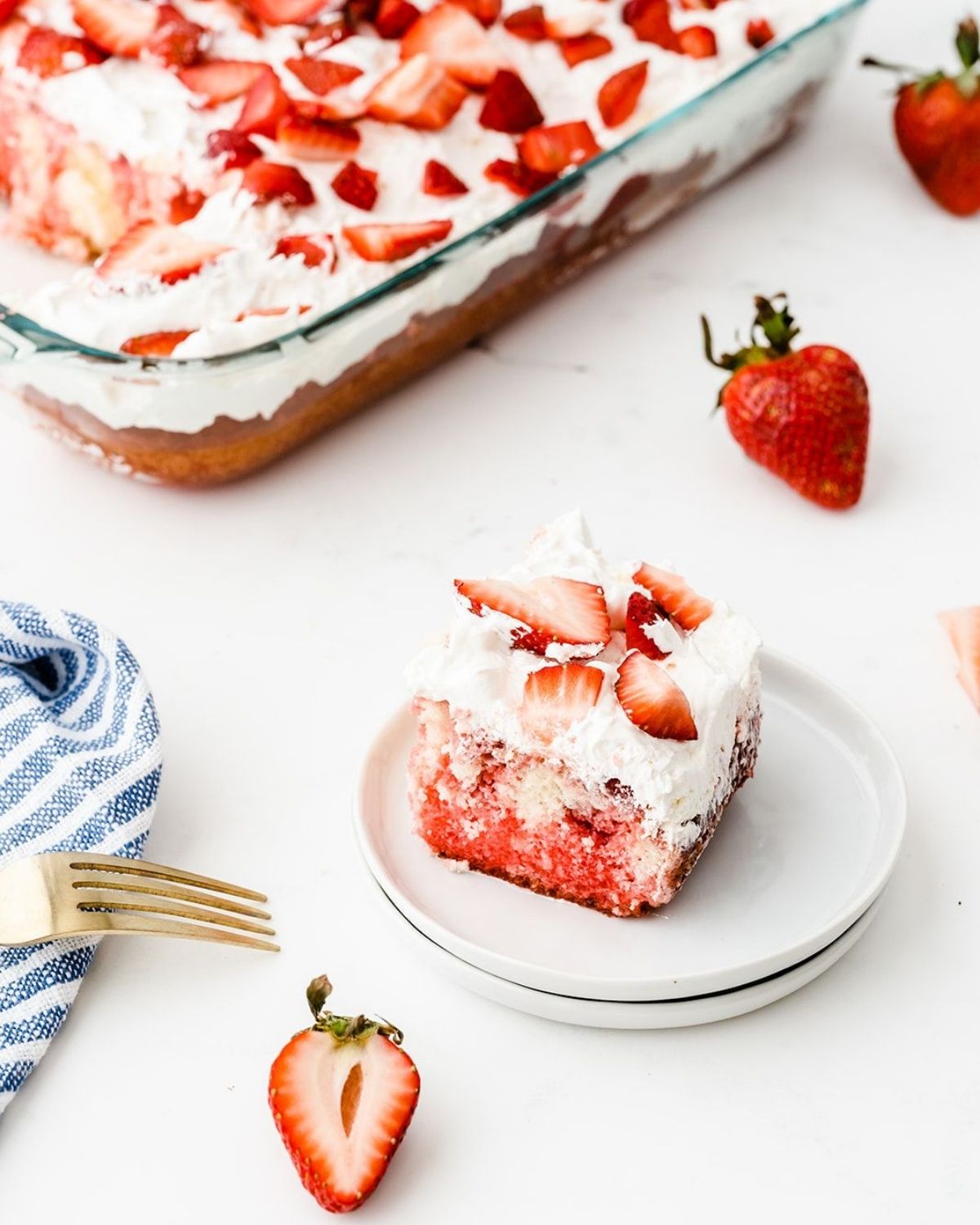 a slice of poke cake strawberry on a stack of white plates with more cake in a glass dish in the background