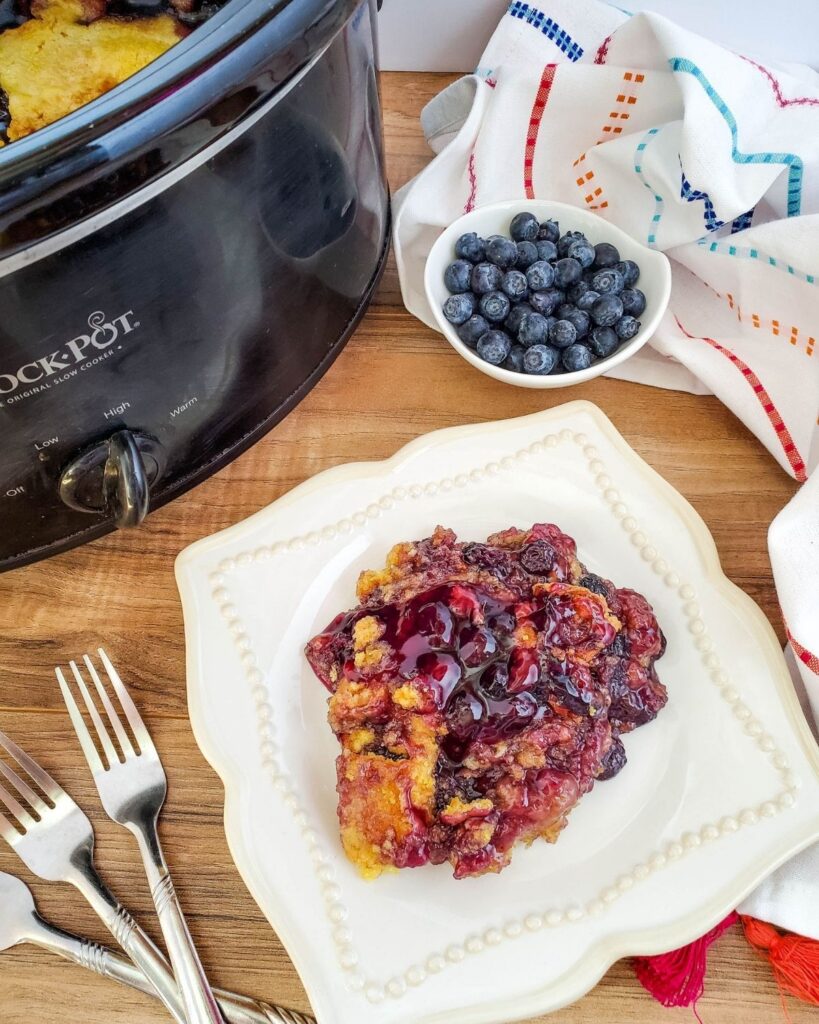 blueberry crockpot cake on a white plate next to forks a bowl of blueberries and a crockpot