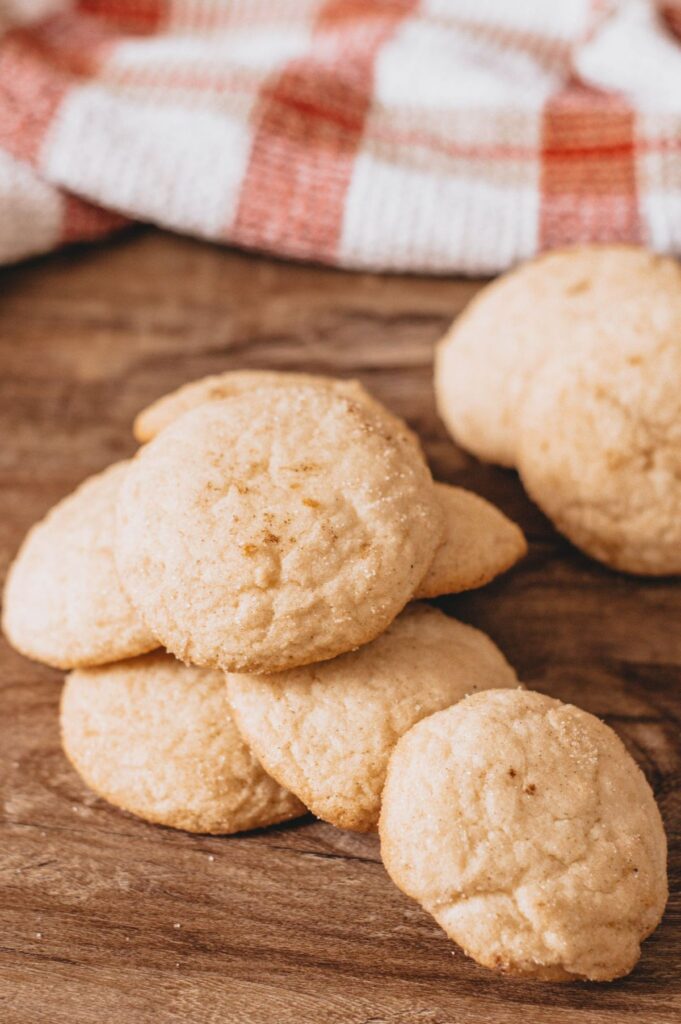 a stack of snickerdoodle cookies on a wood table with more cookies and a red and white cloth blurred in the background