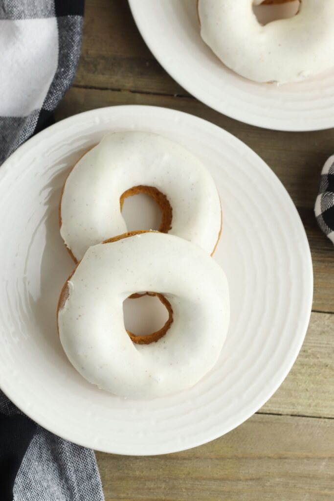 a white plate with 2 spiced pumpkin donuts on it with another donut on a plate in the background
