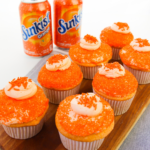 7 sunkist orange cupcakes on a platter next to two cans of sunkist soda
