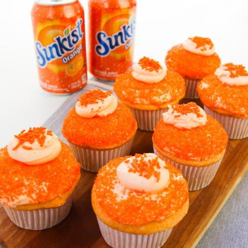 7 sunkist orange cupcakes on a platter next to two cans of sunkist soda