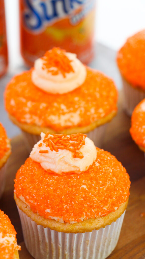 a closeup of 2 creamsicle cupcakes with sunkist soda cans in the background