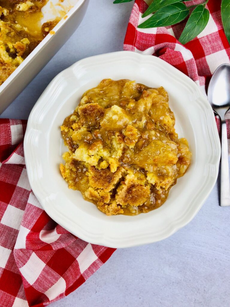 apple dump cake with caramel on a white plate next to a spoon on a red and white checkered cloth and the rest of the cake in a pan