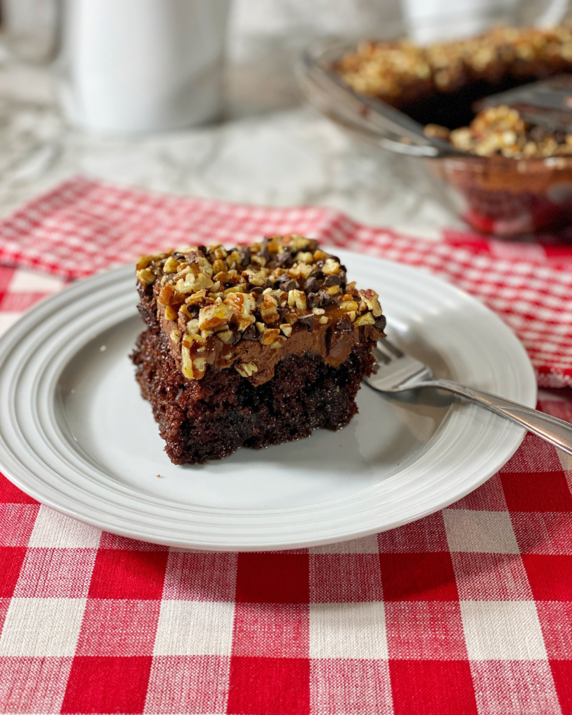 a slice of caramel chocolate Poke Cake topped with nuts on a plate with a fork on the side.