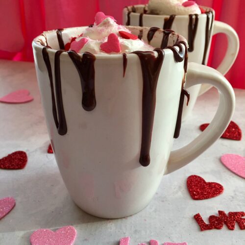 two white mugs filled with hot chocolate mug cake, topped with cool whip, fudge drizzle and heart shaped sprinkles