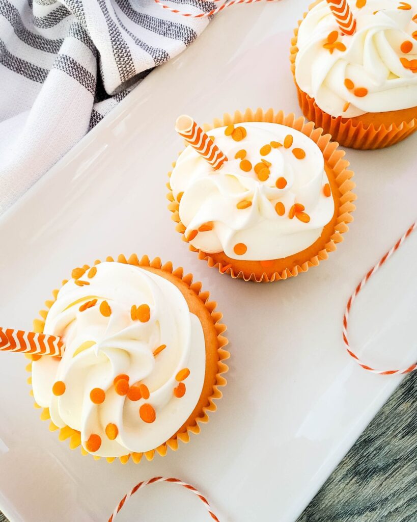 overhead view of 3 creamsicle orange cupcakes on a white platter next to a gray and white cloth
