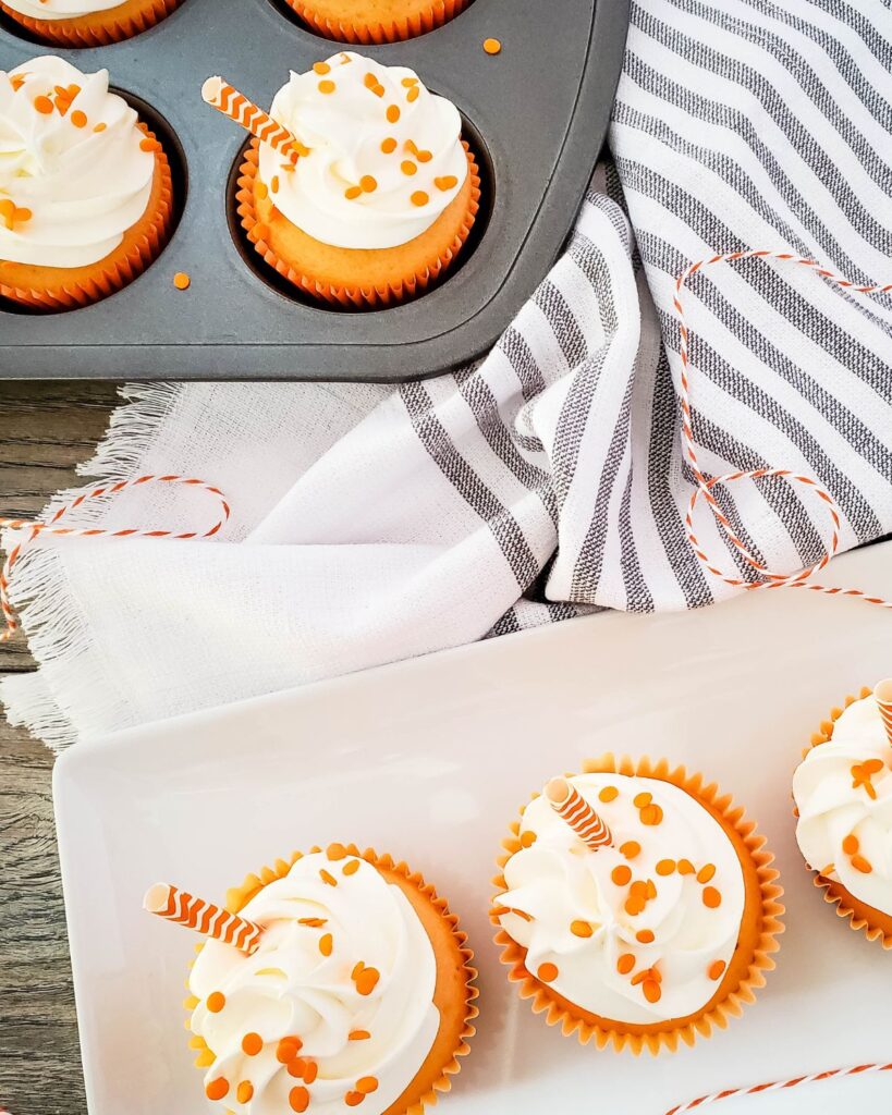 3 creamsicle orange cupcakes on a white platter with more cupcakes in a pan in the background