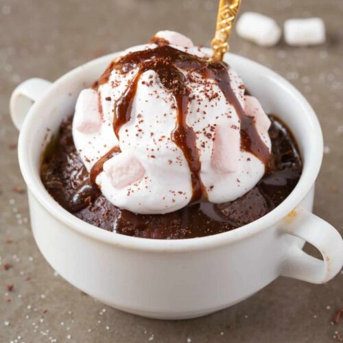 a big white mug of hot chocolate mug cake topped with cool whip and fudge drizzle with a gold spoon in the side.