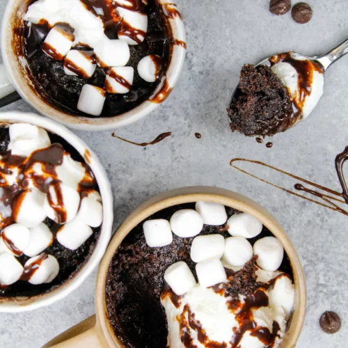 three hot chocolate mug cakes topped with white mini marshmallows and chocolate fudge drizzle.