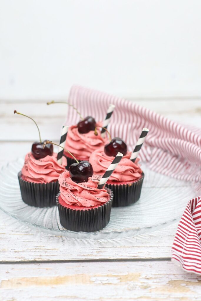 4 Cherry Cupcakes with a fresh cherry on top  and  a straw on the side on a glass plate next to a red and white striped cloth