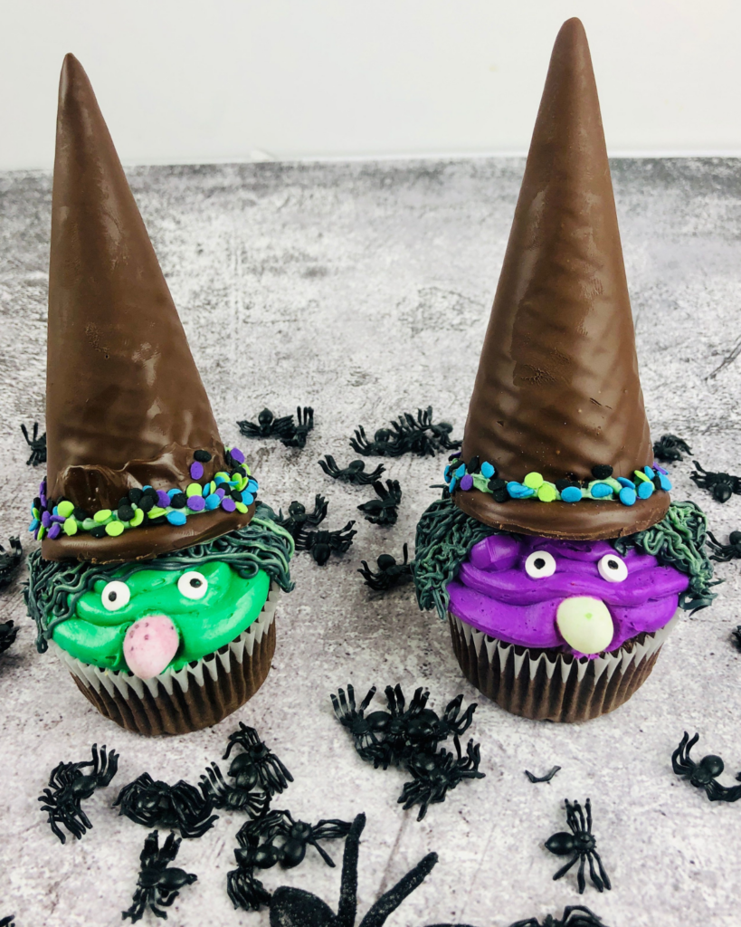 two Witch Cupcakes one green and one purple with plastic spiders in the background.
