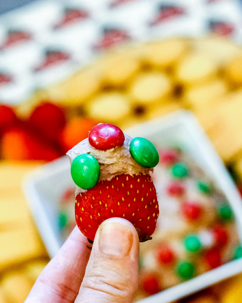 a closeup of a hand holding a Strawberry dipped in Brownie Batter Dip with m&m's on top. 