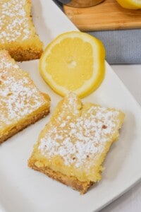 Two lemon bars on a plate with a slice of lemon on the side.